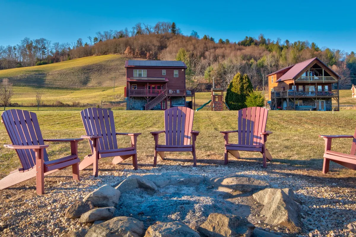A cozy outdoor gathering area with adirondack chairs arranged around a large fire pit, set against wooden houses and lush green hills, evoking relaxed evenings and group get-togethers.
