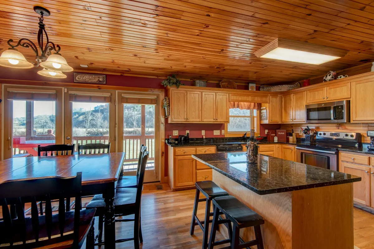 A modern kitchen and dining area with a granite kitchen island, wooden cabinets, and a large dining table. Expansive windows offer picturesque views of the surrounding landscape, filling the space with light and emphasizing the open-plan design.