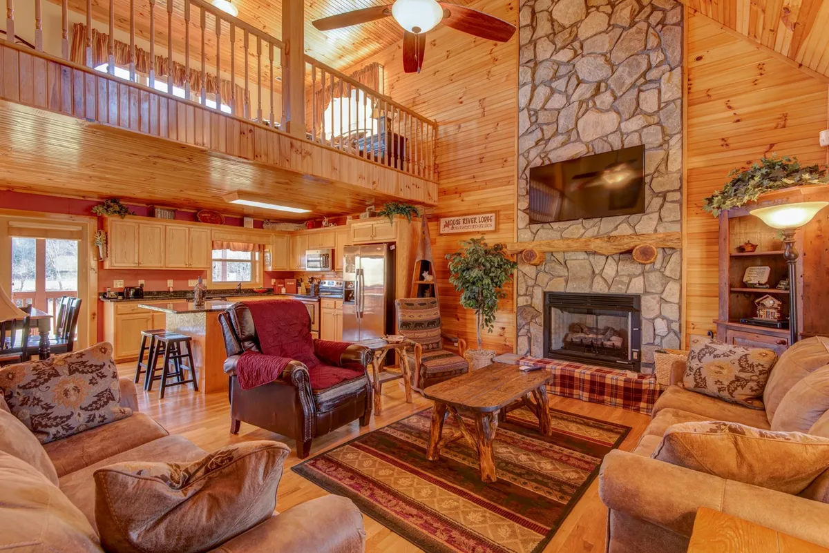 A spacious open-plan interior featuring a striking floor-to-ceiling stone fireplace, wooden ceiling beams, comfortable leather seating, and a kitchen area in the background. Large windows allow natural light to flood the space and offer views of the outdoors, while the layout invites relaxation and togetherness.