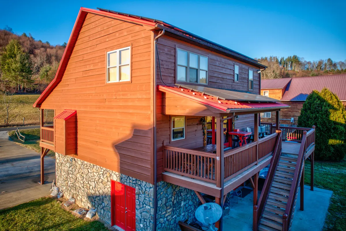 This image features the exterior of the property, showcasing wooden siding, a stone foundation, and a welcoming red door. The outdoor seating area on the porch is visible, suggesting a comfortable spot to relax and take in the mountain air. The overall setting feels cozy and inviting, perfect for couples seeking privacy and tranquil surroundings.