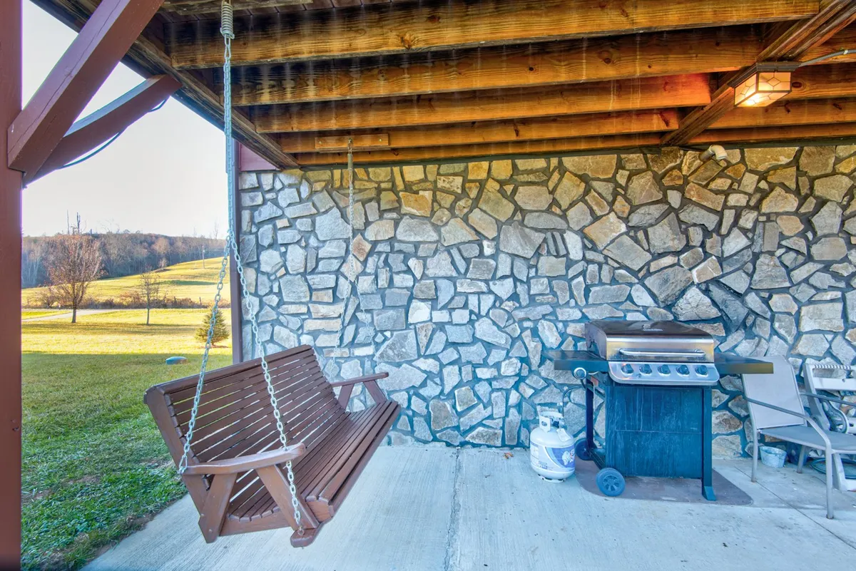 This image features a welcoming outdoor space with a wooden swing, stone wall, grill, and comfortable seating. The scene is relaxed and inviting, suggesting an ideal spot for barbecues, socializing, or simply unwinding while surrounded by nature.