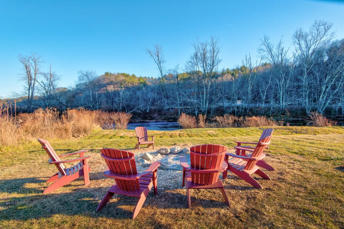 This image features red Adirondack chairs arranged around a fire pit on a grassy area, with a striking view of the river in the background. The evening light highlights the tranquil riverfront setting, and the arrangement clearly invites gatherings for storytelling, relaxation, and stargazing.
