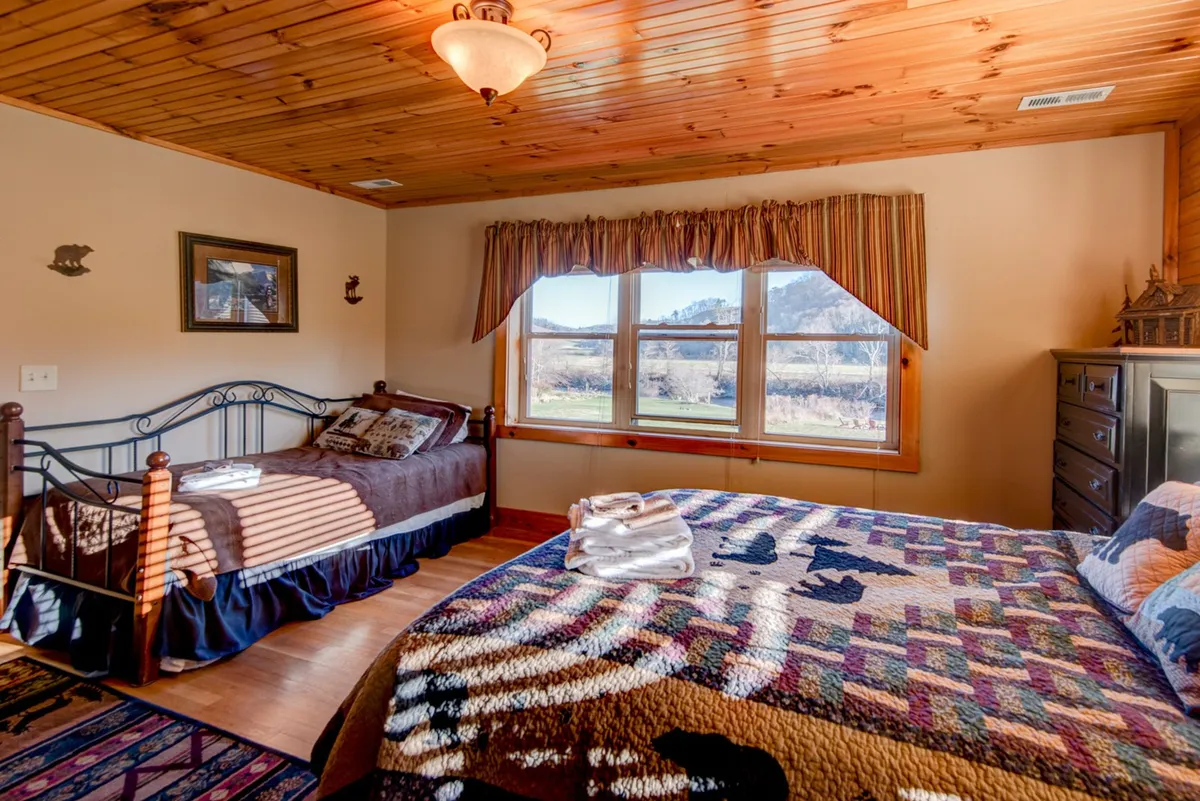 A lofted space featuring two beds beneath a wooden ceiling, a large window framing a scenic landscape, and a relaxed, homey atmosphere. The arrangement suggests privacy and is ideal for families or groups seeking a quiet retreat.