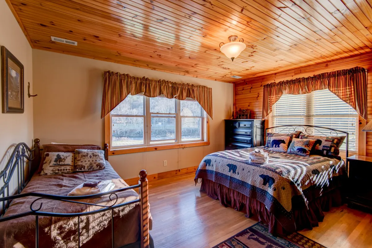 A spacious bedroom with a large window letting in ample natural light, decorative bedding featuring bear motifs, a rustic wooden ceiling and flooring, and a tranquil sitting area by the window. The view outside hints at river scenery, and the overall atmosphere is cozy yet refined, blending rustic charm with comfort.