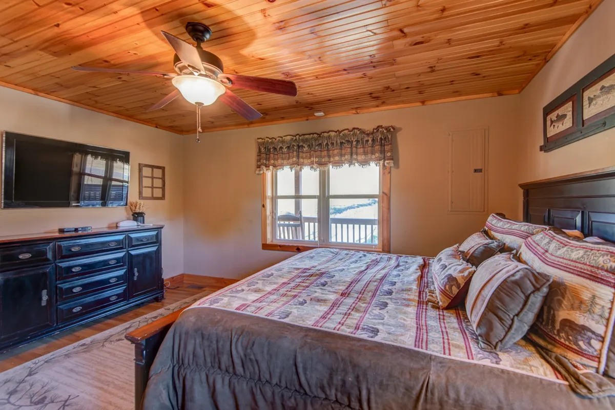 A queen-sized bed with decorative pillows, a wooden ceiling fan, a television mounted on the wall, a window with curtains that allows in natural light, and a dark wooden dresser, creating a warm and inviting bedroom retreat.