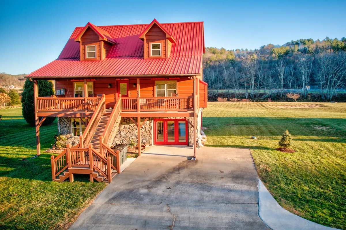 The image showcases a charming cabin with a striking red metal roof, nestled on a stone foundation. Wooden stairs lead up to a spacious porch with large windows that invite natural light inside. The cozy exterior is surrounded by lush greenery, evoking a sense of seclusion and warmth—perfect for gatherings, barbecues, and relaxing while watching sunsets or winter snowfalls.