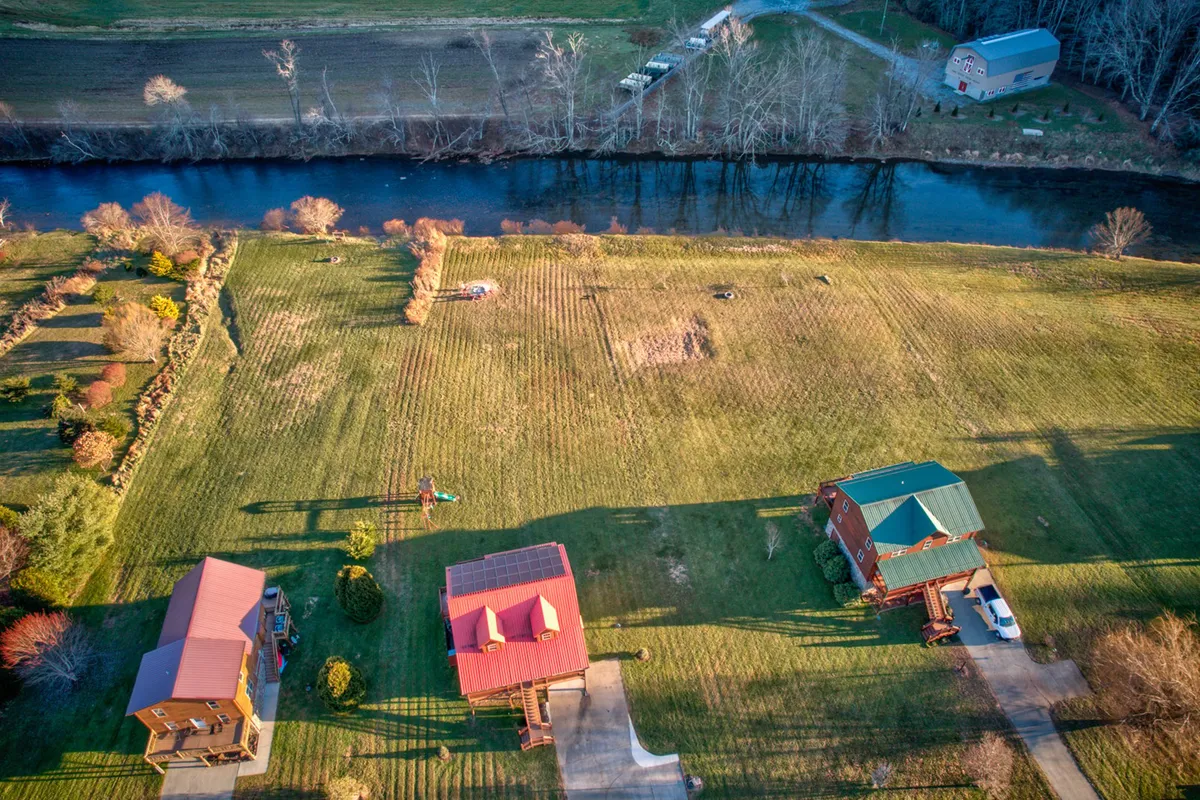 This image presents three distinct houses set along the riverbank, with manicured gardens and inviting lawns, all framed by the flowing river and surrounding landscape. The scene conveys a sense of community, spaciousness, and the variety of activities possible in the outdoor environment, from fishing to picnicking by the water’s edge.