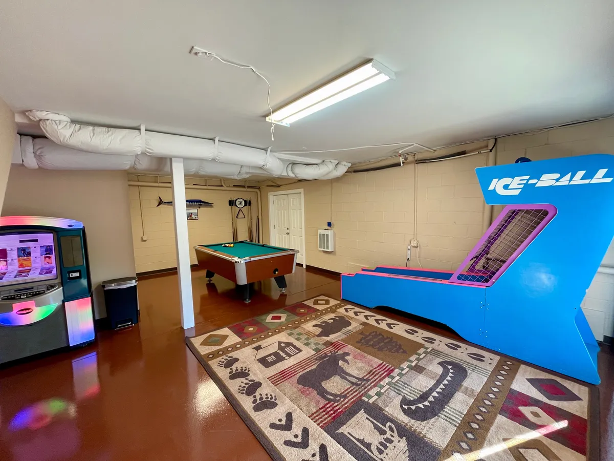 An energetic game room featuring a pool table with animal motif carpeting, a vibrant arcade game machine, and a vending machine. The space is lively, colorful, and clearly designed for fun group activities and friendly competition.