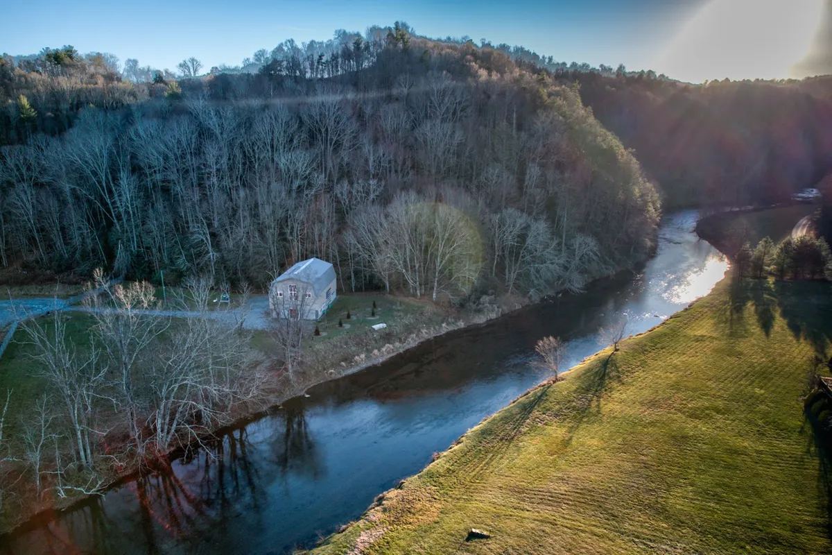 This image features the house situated alongside the New River, surrounded by lush trees and natural landscaping. The tranquil river setting is clearly visible, inviting thoughts of fishing, kayaking, or simply relaxing by the water. The presence of open space and greenery suggests immediate access to outdoor activities and the beauty of the Blue Ridge region.